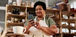 Low angle portrait of African American senior woman using smartphone in pottery studio and posting pictures on social media copy space