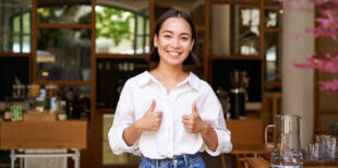 Happy asian woman shows thumbs up, approves, likes smth good, stands near cafe entrance and give her recommendation.
