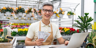 A smiling Caucasian man holds money, works on a laptop at a flower shop, surrounded by beautiful plants.