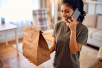 Hispanic woman talking on cell phone after receiving home delivery in paper bag.