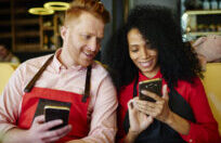African American waitress sitting next to male colleague on sofa and showing phone screen while using gadgets and chilling during break
